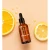 Yokaba Orange Oil 50 ml na brzoskwiniowym tle między plastrami pomarańczy, obok krople olejku