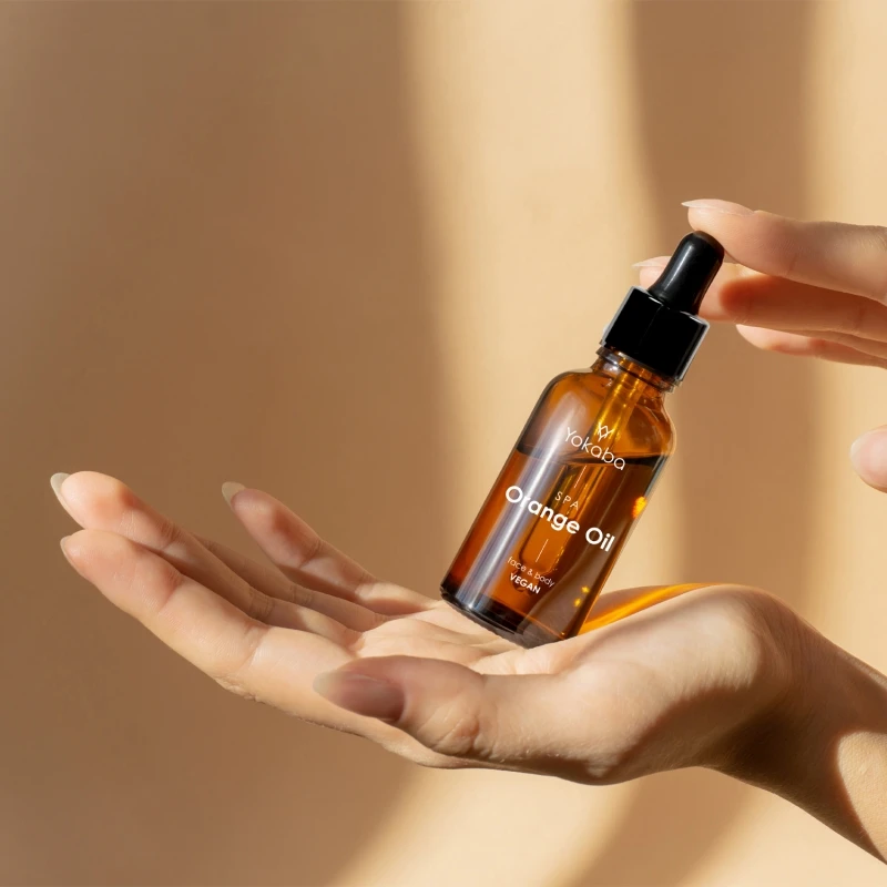Dłonie podające butelkę olejku Yokaba Orange Oil 50 ml w ciepłym, słonecznym świetle